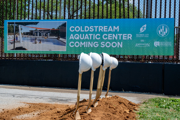 Dirt with shovels sticking up in it with white hard hats on top of the shovel handles.  Banner in background announcing Coldstream Aquatic Center coming soon.